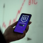 Mão segurando um smartphone com um aplicativo de blockchain, diante de um gráfico de ações ao fundo.