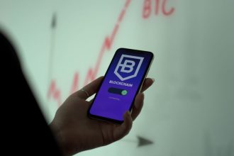 Mão segurando um smartphone com um aplicativo de blockchain, diante de um gráfico de ações ao fundo.