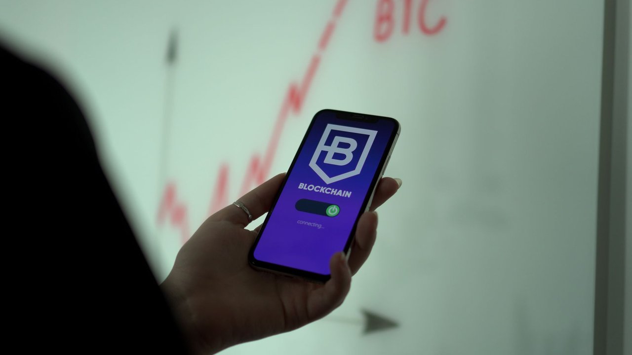 Mão segurando um smartphone com um aplicativo de blockchain, diante de um gráfico de ações ao fundo.