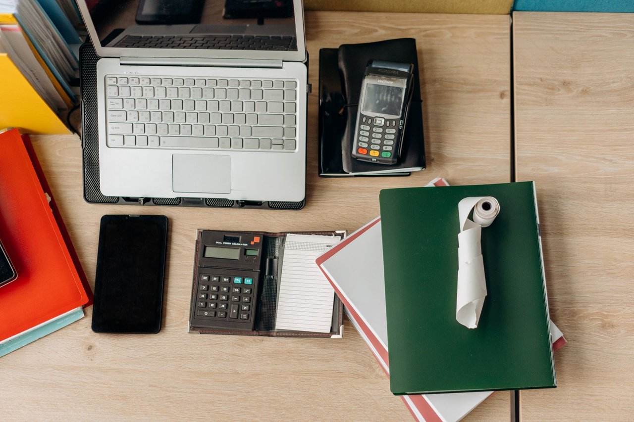 Mesa de escritório organizada com laptop, calculadora, terminal de pagamento e documentos.