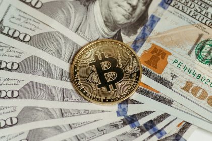 Moeda de Bitcoin sobre uma pilha de notas de cem dólares.