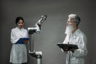 Dois pesquisadores analisam um braço robótico em um laboratório de tecnologia.